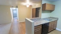 This single-story space is designed with an open-concept layout that connects the kitchen and living area, creating a cohesive and functional environment. The kitchen features a peninsula with granite-style laminate countertops, dark cabinetry providing generous storage, and a black dishwasher integrated seamlessly beneath the counter. Upper cabinetry in a warm brown finish adds additional storage and frames the cooking space. The peninsula serves as a natural divider between the kitchen and the adjoining living area, making it ideal for casual dining or entertaining.

The living area is finished with warm hardwood-style flooring that extends throughout, adding continuity and a sense of warmth to the space. A ceiling fan with an integrated light fixture is centrally positioned, offering both air circulation and ambient lighting. A spacious built-in closet with a sliding door and interior hanging rods provides notable storage capacity within the living area.

A hallway visible beyond the living space leads further into the unit, suggesting additional rooms. Natural light filters in from windows visible down the corridor, contributing to a bright and welcoming overall ambiance throughout this practical, well-organized space.