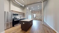 Located at 150 N 3rd St in the heart of downtown Columbus, Ohio, this single-floor urban loft masterfully blends historic character with modern finishes. The open-plan layout is anchored by warm hardwood flooring that flows seamlessly throughout the entire space, complemented by soaring ceilings that enhance the sense of volume and openness.

The kitchen is a standout feature, equipped with a full suite of stainless steel appliances including a gas range, microwave, refrigerator, and dishwasher. A central island with a crisp white quartz countertop serves as the focal point, while open wood shelving and a classic white subway tile backsplash add contemporary style. Exposed industrial ductwork overhead reinforces the loft's urban aesthetic.

The living area is framed by a striking exposed brick wall — partially whitewashed to reveal layers of history — punctuated by an arched window that frames views of the surrounding cityscape. Original structural columns add to the authentic industrial character of the space.

A bedroom area includes open closet shelving and direct sightlines to the main living space. The bathroom features hardwood-style flooring, a modern vanity with black fixtures, and clean, neutral finishes. Throughout, the combination of raw architectural elements and refined modern updates creates a cohesive, sophisticated urban environment.