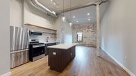 Located at 150 N 3rd St in the heart of downtown Columbus, Ohio, this single-floor urban loft masterfully blends historic character with modern finishes. The open-plan layout is anchored by warm hardwood flooring that flows seamlessly throughout the entire space, complemented by soaring ceilings that enhance the sense of volume and openness.

The kitchen is a standout feature, equipped with a full suite of stainless steel appliances including a gas range, microwave, refrigerator, and dishwasher. A central island with a crisp white quartz countertop serves as the focal point, while open wood shelving and a classic white subway tile backsplash add contemporary style. Exposed industrial ductwork overhead reinforces the loft's urban aesthetic.

The living area is framed by a striking exposed brick wall — partially whitewashed to reveal layers of history — punctuated by an arched window that frames views of the surrounding cityscape. Original structural columns add to the authentic industrial character of the space.

A bedroom area includes open closet shelving and direct sightlines to the main living space. The bathroom features hardwood-style flooring, a modern vanity with black fixtures, and clean, neutral finishes. Throughout, the combination of raw architectural elements and refined modern updates creates a cohesive, sophisticated urban environment.
