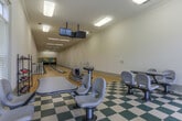 Located at 8064 S Fulton Pkwy in Fairburn, Georgia, this single-floor bowling facility offers a well-appointed recreational experience. The space transitions seamlessly from a casual seating and lounge area into the dedicated bowling lanes, creating a cohesive and functional layout throughout.

The lounge area features a distinctive green-and-white checkered tile floor that lends the space a classic, retro character. Multiple wall-mounted flat-screen televisions are positioned for easy viewing from the seating areas, which include pedestal-style tables and chairs suited for groups of various sizes. Large windows dressed with horizontal blinds line the walls, allowing generous amounts of natural light to brighten the interior while offering views of the surrounding greenery.

A dedicated rack holds bowling shoes in multiple sizes, conveniently positioned near the transition point between the lounge and the lanes. The bowling area itself features two full-length lanes finished in warm hardwood, extending toward vibrant pin artwork at the far end. Overhead, an additional cluster of monitors hangs above the lanes for score tracking and entertainment. The lanes are framed by clean white walls adorned with crown molding details, and the ceiling height gives the space an open, airy feel. The overall ambiance is bright, welcoming, and well-suited for recreational use.
