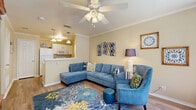 This cozy yet thoughtfully designed single-floor unit offers a warm and inviting atmosphere highlighted by consistent wood-look flooring, crown molding, and a cohesive blue-and-yellow color scheme throughout. The open living area is anchored by a ceiling fan with light fixture, large windows that invite ample natural light, and a bay window nook that accommodates a small dining table — perfect for everyday meals. The living space flows comfortably into a compact galley-style kitchen featuring white cabinetry with gold hardware, white appliances, a double sink, and a pass-through that maintains visual connectivity with the living area. The bedroom is a comfortable retreat with dual windows dressed in navy curtains, a ceiling fan, crown molding, and hardwood-style flooring. A wooden dresser with mirror provides generous storage, and the room benefits from natural light on multiple sides. The full bathroom is neatly appointed with a white vanity, a tiled shower-tub combination, and wood-look flooring. A dedicated closet provides additional storage. Overall, the space is efficiently laid out, making excellent use of every square foot with a bright, cheerful character.