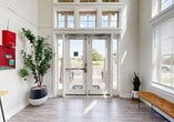 This well-appointed community clubhouse offers a thoughtfully designed single-floor layout encompassing a generous amount of functional and social space. The grand entrance immediately impresses with soaring double-height ceilings, floor-to-ceiling glass double doors framed by transom windows that flood the foyer with natural light, and light wood-style flooring that flows throughout. Lush potted greenery and a bench seating area create a welcoming atmosphere upon arrival.

The open lounge area serves as the social heart of the facility, anchored by a dramatic floor-to-ceiling stacked stone fireplace with a wood mantle and wall-mounted television. Rich leather seating, a round wood coffee table, and playful wall décor featuring vintage propeller accents give the space a warm, inviting character with soaring ceilings overhead.

The kitchen is fully equipped for entertaining and everyday use, featuring granite countertops, dark cabinetry, stainless steel appliances including a full refrigerator, dishwasher, and microwave, plus a beverage station with generous counter space and abundant natural light from large windows.

The fitness center is spacious and well-lit, outfitted with treadmills, rowing machines, stationary bikes, free weights, and multi-use strength equipment, with large windows and garage-style doors providing outdoor views and ventilation.