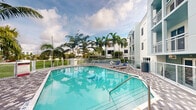 Located at 151 Southeast 3rd Avenue in Delray Beach, Florida, this outdoor amenity space offers a quintessential South Florida poolside experience. The centerpiece is a generously sized, freeform swimming pool with crystal-clear turquoise water, framed by a patterned tile deck that adds a refined aesthetic to the area. Blue lounge chairs are positioned poolside, providing a dedicated space for relaxation and sun-soaking. The surrounding landscape features several tall, mature palm trees that create a lush tropical atmosphere, evoking the natural beauty of the region. A metal safety fence encloses the pool area, ensuring security while maintaining an open, airy feel. The pool is situated directly adjacent to a modern, multi-story residential building with a clean white exterior and balconies, whose contemporary architectural design complements the outdoor setting. Open green space is visible in the background, adding to the sense of openness. The space is exposed to Florida's abundant natural sunlight, with the sky and clouds reflected in the pool's shimmering surface. This amenity area serves as a serene outdoor retreat, well-suited to the warm climate of Delray Beach.
