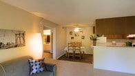 This single-floor residence is designed with an open-concept layout that seamlessly connects the living, dining, and kitchen areas into one cohesive and functional space. The living area is finished with plush carpeting and is anchored by a comfortable seating arrangement accented by decorative throw pillows, complemented by a tall floor lamp that adds a warm ambient glow to the room.

The dining area transitions naturally from the living space, featuring hardwood-style flooring that defines the zone. A set of wooden ladder-back chairs surrounds a dining table beneath a ceiling fan fitted with a light fixture, providing both airflow and illumination. The neutral-toned walls are adorned with a modest selection of framed artwork, adding a personalized touch without overwhelming the space.

Adjacent to the dining area, a kitchen peninsula with light-colored countertops provides a practical divide between the cooking and dining zones. Upper wooden cabinetry is visible, offering ample storage. A hallway visible from the living area leads to what appears to be a bathroom, featuring bright lighting and tiled walls. Overall, the space exudes a cozy, well-lit, and practical atmosphere.
