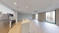 This single-floor space showcases a clean, contemporary design characterized by a neutral palette of whites and soft grays throughout. The open-concept living and kitchen area forms the heart of the unit, anchored by a sleek central island with a light countertop and an integrated sink. The kitchen is equipped with stainless steel appliances, including a refrigerator, electric range, and built-in microwave, all complemented by tall white cabinetry that maximizes vertical storage. Light-toned hardwood-style flooring extends seamlessly across the main living area, enhancing the sense of openness and flow.

The living area is generously proportioned, benefiting from multiple floor-to-ceiling windows with slim black frames that draw in substantial natural light and provide views of the surrounding outdoors. A ceiling-mounted HVAC cassette and recessed lighting ensure year-round comfort and even illumination throughout the space.

Two bathrooms serve the unit. The primary bathroom features large-format white tile flooring, a floating white vanity with an undermount sink, an illuminated mirror, and a glass-enclosed walk-in shower. The secondary bathroom offers a full tub-shower combination with white tile surround, a matching vanity, and consistent clean finishes. Overall, the space presents a bright, move-in-ready environment with a cohesive modern aesthetic.