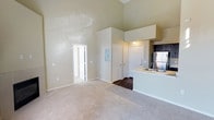 This two-story residence makes an immediate impression with its dramatically high ceilings in the main living area, amplifying the sense of openness and space. The living room is finished with plush wall-to-wall carpeting in a neutral tone, contributing to a warm and comfortable environment. Soft, light-toned walls throughout enhance the brightness of the interior.

The living space flows naturally toward the kitchen area, where the flooring transitions to hardwood-style dark planks, providing a distinct visual contrast. The kitchen features a peninsula with a built-in sink, light-colored countertops, and dark cabinetry, complemented by a stainless steel refrigerator. This layout makes the kitchen both functional and visually connected to the main living space.

Multiple interior doors are visible, suggesting well-defined separate rooms accessible from the main living area. A utility panel is neatly recessed into the wall, maintaining a clean aesthetic. The overall design emphasizes vertical space and an open-concept arrangement, making the residence feel expansive and well-organized across its two floors.