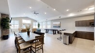 Located on W Stoney Creek Dr in Rogers, Arkansas, this single-floor community clubhouse offers a thoughtfully designed gathering space that blends comfort with functionality. The open layout is anchored by a striking stone-clad fireplace with a mounted flat-screen TV above it, creating an inviting focal point in the lounge area. Plush seating, including an upholstered sofa and accent chairs, surrounds the fireplace, making it an ideal spot for relaxation and conversation.

The space flows seamlessly into a well-equipped kitchenette, outfitted with rich dark wood cabinetry, light granite countertops, a white tile backsplash, stainless steel appliances including a built-in microwave and dishwasher, and a double-basin sink. A peninsula with bar-height seating provides an additional casual dining option.

A generously sized dining area accommodates a large dark wood table with ample seating, suitable for group gatherings. Adjacent to this is a recreation zone featuring a full-length shuffleboard table, flanked by a cozy seating nook with a tufted blue sofa. Large windows throughout flood the space with natural light and offer views of the surrounding community. Light tile flooring, recessed lighting, and a ceiling fan complete the clean, modern aesthetic of this versatile clubhouse.