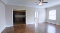 This single-floor residence offers a well-connected layout that balances open living with functional room separation. The main living area is spacious and bright, featuring rich, dark hardwood flooring that extends throughout the space, crisp white crown molding along the ceiling, and a ceiling fan with integrated lighting that adds both comfort and style. Large baseboards provide a clean, classic finish to the walls. An open doorway leads directly into the kitchen, which is outfitted with dark wood cabinetry offering generous storage, a double sink, and dark countertops. A full-size refrigerator anchors one end of the kitchen, while a dishwasher completes the practical layout. The kitchen flooring transitions to a complementary tone, subtly distinguishing the cooking space from the living area. From the living room, a second doorway opens to an additional room that benefits from natural light, hinting at versatility as a bedroom or den. The overall ambiance of the space is neutral and inviting, with light gray walls providing a clean canvas throughout. The combination of classic architectural detailing and warm wood tones gives the residence a timeless and comfortable character.