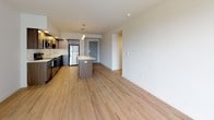 This single-floor unit offers a thoughtfully designed open-concept layout that seamlessly connects the kitchen, dining, and living areas. The kitchen is a standout feature, equipped with stainless steel appliances including a refrigerator, electric range, and microwave. Rich, dark wood cabinetry lines the walls from floor to ceiling, complemented by crisp white quartz countertops and a central island that doubles as a breakfast bar, illuminated by stylish pendant lighting overhead. Light wood-tone flooring flows throughout the main living areas, enhancing the bright, airy atmosphere. The living area is generously proportioned and benefits from large floor-to-ceiling windows and glass doors that open to a balcony, flooding the space with natural light and providing outdoor views. The bedroom features soft wall-to-wall carpeting and clean white walls, offering a calm and neutral retreat. The bathroom is finished with light wood-tone flooring, a vanity with dark wood cabinetry, and a full tub-shower combination with chrome fixtures. Throughout the unit, the clean lines, neutral color palette, and modern finishes contribute to a polished, contemporary aesthetic that is both functional and inviting.