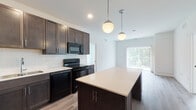 Located at 27W555 High Lake Rd in Winfield, Illinois, this single-floor residence offers a thoughtfully designed layout with contemporary finishes throughout. The kitchen serves as a central highlight, showcasing rich dark-stained cabinetry extending to the ceiling, light quartz countertops, a white subway tile backsplash, and a full suite of black appliances including a refrigerator, range, built-in microwave, and dishwasher. A substantial kitchen island with matching cabinetry and light countertop provides additional workspace and seating potential. Pendant globe lighting overhead adds a polished, modern touch. The kitchen flows naturally into the living area, which features light wood-style flooring and a sliding glass door with vertical blinds that floods the space with natural light and provides outdoor access. Bedrooms are finished with neutral carpeting and large windows that frame outdoor greenery, lending each room a calm, airy atmosphere. The bathrooms are clean and functional, with one featuring a spacious walk-in shower enclosed by a framed glass door with white tile surround, and another featuring a dark wood vanity with a white quartz countertop and a large mirror.