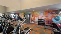 This fitness center is a thoughtfully designed, single-floor workout facility that combines functionality with an energizing aesthetic. The space is anchored by a striking full-wall mural featuring a tropical motif with the motivational phrase "Stronger Than Yesterday," rendered in bold orange, blue, and yellow tones that set an uplifting atmosphere throughout the room. The geometric patterned carpet — in vivid orange and gray — adds a dynamic visual energy underfoot and complements the mural's color palette.

The cardio zone is well-stocked with multiple treadmills, elliptical cross-trainers, and a stationary bike, all arranged to take advantage of the natural light streaming in through several windows that offer views of the surrounding greenery. A ceiling fan overhead provides additional airflow and comfort during workouts.

On the strength training side, a multi-station cable machine provides versatile resistance options, while a full dumbbell rack — ranging across a variety of weights — supports free weight training. Stability balls are also available for functional fitness exercises.

Full-length mirrors line key walls, amplifying the sense of space and allowing users to monitor their form. Recessed lighting throughout ensures the room is consistently well-lit. The high, angled ceilings contribute to an open, airy feel, making the fitness center feel spacious and welcoming.