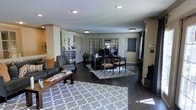 This single-floor space is designed to function as a professional office environment with a warm, residential aesthetic. The open layout seamlessly integrates a reception or lounge area with a central workspace, creating a fluid and functional floor plan. Dark hardwood floors run throughout, grounding the space and providing a rich contrast against the neutral beige and white wall tones. Recessed and flush-mount ceiling lighting ensures consistent illumination across the space, complemented by natural light streaming in through a front-facing door with sidelight windows. The workspace area features a substantial dark wood desk with computer equipment and guest seating, anchored by a geometric-patterned area rug. A matching rug defines the lounge zone, which includes upholstered gray armchairs and a coffee table with reading materials — ideal for client reception. Decorative grid-pane mirrors on the back wall add depth and a stylish architectural element. A promotional display banner and interior plants contribute to a polished, inviting atmosphere. Crown molding and a textured ceiling add refined finishing touches throughout.