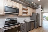 This single-floor unit offers a thoughtfully designed open layout that blends modern finishes with industrial character. The kitchen is efficiently arranged along a single wall and features stainless steel appliances, including an electric range, over-the-range microwave, dishwasher, and refrigerator. Light natural wood cabinetry on the upper portion contrasts with darker lower cabinets, while clean white countertops provide ample prep space. Exposed metallic ductwork running overhead adds a distinctive industrial aesthetic throughout the unit.

The open living area is spacious and bright, with light-toned hardwood-style flooring that extends throughout the entire space. A glass-paneled door leads to a private balcony, allowing natural light to fill the room and offering an outdoor connection. Dark baseboards provide a clean contrast against the neutral white walls.

The bathroom is well-appointed with a walk-in glass-enclosed shower, a vanity with an integrated sink, a large mirror with overhead lighting, and a toilet. Consistent hardwood-style flooring ties the bathroom seamlessly to the rest of the unit.

An electrical panel is built into one wall, and the exposed HVAC ductwork remains a prominent architectural feature, giving the space an urban loft-inspired character throughout.
