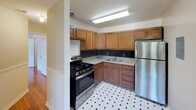 This single-floor space is defined by its consistent use of warm, polished hardwood flooring that runs through the living and bedroom areas, lending a cohesive and well-maintained character to the interior. The living area is generously proportioned, featuring neutral-toned walls, white baseboard trim, and two closeted storage areas accessible via white panel doors. An adjacent room, suitable as a bedroom, includes a ceiling fan with integrated lighting, a window fitted with blinds for natural light control, and a radiator heating unit beneath the sill. A second similarly sized room also features a radiator and a window offering outdoor views, complemented by a flush-mount ceiling light fixture. The kitchen is a functional, well-equipped space with warm medium-toned wood cabinetry arranged in an L-shape, a dark mosaic tile backsplash, light granite-style countertops, stainless steel gas range, and a stainless steel refrigerator. Classic black-and-white geometric tile flooring adds visual interest. The bathroom includes a white vanity with an integrated sink, a mirrored medicine cabinet with built-in lighting, a toilet, and a tub-shower combination enclosed with sliding glass doors.