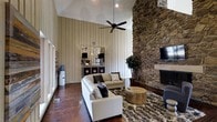 This single-floor space makes a bold architectural statement with its soaring vaulted ceilings that amplify the sense of openness and grandeur throughout. The living area is anchored by a commanding floor-to-ceiling natural stone fireplace, which serves as the room's defining focal point and adds a rugged, organic texture to the interior. Opposite the fireplace, vertical shiplap wall paneling in a crisp, neutral tone provides a clean, contemporary contrast to the stonework, lending the space a refined yet relaxed character. Rich, dark hardwood flooring runs throughout, grounding the room with warmth and depth. A large ceiling fan with dark-finished blades hangs centrally from the vaulted ceiling, offering both function and a visual complement to the overall design. The living area is furnished with plush, neutral-toned seating arranged around a textured area rug and a rustic wooden coffee table, creating an inviting and comfortable gathering space. Recessed lighting supplements the natural light that filters in through large windows, ensuring the space is well-lit at any time of day. A mounted flat-screen television is positioned above the fireplace mantle, seamlessly integrating modern amenities into the classic, nature-inspired design. Overall, the space strikes a balanced blend of rustic charm and contemporary sophistication.
