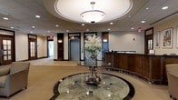 This sophisticated lobby space makes an immediate impression with its refined architectural design and upscale finishes. At the heart of the entrance is a striking rotunda, defined by a recessed circular ceiling detail housing a large, elegant pendant light fixture that bathes the space in warm, ambient illumination. The floors feature polished marble with a circular inlay pattern, reinforcing the grandeur of the rotunda and anchoring a central ornate pedestal table adorned with an elaborate floral arrangement, adding a touch of classic elegance. Recessed lighting throughout the ceiling complements the main fixture, ensuring the space is well-lit and inviting. A reception desk finished in rich, dark wood sits to one side, providing a professional and welcoming point of service for visitors. Dark wood paneling and trim accents frame the elevator bays, contributing to the space's distinguished aesthetic. The flooring transitions from polished marble in the central rotunda area to a neutral carpeted corridor, guiding visitors seamlessly toward additional areas of the building. Overall, the lobby conveys a sense of prestige and professionalism, combining classic architectural elements with carefully chosen decorative details to create a formal and welcoming environment.
