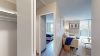 This single-floor residence offers a thoughtfully designed layout that balances comfort and functionality. The living room serves as the heart of the unit, featuring wood-look flooring, neutral tones, and a large sliding glass door fitted with vertical blinds that welcomes an abundance of natural light and provides access to an outdoor area. The space is well-proportioned, comfortably accommodating a sofa, area rug, and coffee table. Adjacent to the living area, the kitchen is equipped with warm honey-toned cabinetry, granite-style countertops, a stainless steel sink, and a black gas range with overhead ventilation — providing ample storage and a practical workspace. A dining area with seating for multiple guests sits conveniently nearby. The bedroom is a serene retreat, highlighted by a large window with garden views, mid-century modern furnishings, and soft, neutral wall tones that create a calming atmosphere. The bathroom features a combined tub and shower with a curtain enclosure, a wood-cabinet vanity with an integrated sink, and a well-lit mirror — finished with the same durable wood-look flooring found throughout the residence. Overall, the space presents a clean, modern aesthetic with warm accents throughout.