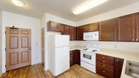 This single-floor unit offers a practical and comfortable layout designed for everyday living. The kitchen serves as a central highlight, featuring rich dark-wood cabinetry that extends to the upper walls for generous storage, light-toned countertops, and a full suite of white appliances including a refrigerator, electric range, over-the-range microwave, dishwasher, and double-basin sink. Warm hardwood-style flooring runs throughout the kitchen and extends into the adjacent living area, creating a cohesive and easy-to-maintain surface. The open transition between the kitchen and the main living space allows natural light from multiple windows to flow freely, giving the area a bright and airy feel. Baseboard heating units are present throughout, ensuring warmth during cooler months. The bedroom is generously sized and features plush carpeting, multiple windows fitted with blinds that invite abundant sunlight, and a ceiling fan for added comfort. The bathroom is clean and straightforward, offering a full-size soaking tub with an overhead shower, white tile surround, and a toilet, all finished in a crisp, neutral palette. White walls throughout the unit provide a fresh, move-in-ready canvas.