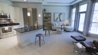 This single-floor residence showcases a thoughtfully designed open-concept layout that seamlessly blends the kitchen and living areas into one cohesive, inviting space. The kitchen features a sleek dark granite countertop bar with modern pendant lighting hanging above, creating an ideal spot for casual dining or entertaining. Industrial-style metal barstools complement the contemporary aesthetic, while a large mirror behind the counter adds depth and brightness to the space.

The living area is generously proportioned, with soft neutral carpeting that adds warmth underfoot and flows naturally from the tiled entryway. A light-toned sectional sofa anchors the seating area, accented by decorative throw pillows and a rounded white coffee table. A standout piece of abstract wall art adds a creative focal point to the room, while a freestanding bookshelf provides both storage and decorative display space.

Large windows dressed with blinds allow natural light to fill the interior, enhancing the soft, cool-toned palette of the walls. Tray ceiling detailing adds architectural character overhead, elevating the overall sense of refinement. The space strikes a balance between modern design and comfortable livability, making it both functional and visually appealing.
