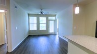 This single-floor residence offers a clean, modern layout designed for comfortable everyday living. The main living area is immediately welcoming, showcasing rich, dark hardwood-style flooring that runs throughout and provides a striking contrast against the light, neutral-toned walls. A ceiling fan with integrated lighting is centrally positioned overhead, contributing to both comfort and style.

Natural light fills the space generously, courtesy of a set of large windows fitted with blinds and a glass-paneled entry door that overlooks the outdoors. The greenery visible through the windows adds a refreshing, connected-to-nature feel to the interior ambiance.

The open-concept design allows the living area to flow seamlessly into the kitchen, which is partially visible from the main space. A countertop with a granite-style finish marks the transition between the two areas, hinting at a functional and well-appointed kitchen space.

The overall layout is thoughtfully designed, with clean lines, ample floor space, and a bright, airy atmosphere. The combination of dark flooring, white trim, and light walls creates a timeless aesthetic that is both inviting and easy to personalize. This space presents a well-balanced environment suited for a variety of living arrangements.