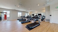 This two-story fitness center is thoughtfully designed to accommodate a wide range of workout needs across its spacious, well-appointed layout. The main level features a dedicated cardio zone equipped with multiple treadmills, ellipticals, and a StairMaster, all arranged on light hardwood flooring with ceiling fans and recessed lighting overhead. Large windows along the perimeter flood the space with natural light and provide views of the surrounding outdoor scenery. Full-length wall mirrors visually expand the area while allowing users to monitor their form. A separate strength training section houses a comprehensive selection of dumbbells, weight benches, cable machines, and functional training equipment, also lined with mirrors for a professional gym atmosphere. A second area on this level incorporates spin bikes, BOSU balls, step platforms, and additional free weights, set beneath soaring double-height ceilings with a striking decorative pendant fixture. Tall, grid-pane windows reach up the full height of this voluminous space, bringing in abundant light. A modern staircase with warm wood treads and sleek glass-panel railings leads to the upper level, where an open mezzanine overlooks the studio below. The overall design balances a clean, contemporary aesthetic with practical, well-equipped functionality.