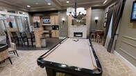 This single-floor entertainment lounge is thoughtfully designed to serve as a premier social and recreational space. At its center sits a full-size billiards table with dark wood rails and neutral felt, positioned beneath an elegant chandelier that anchors the room with warm, focused lighting. Wall sconces throughout the space complement the ambient light, contributing to a refined yet relaxed atmosphere.

A linear fireplace with a classic white mantel serves as a focal point along one wall, adorned with decorative accents that add a polished touch. Adjacent to the fireplace, a high-top bar seating area with stools provides an intimate spot for conversation.

On the opposite side, a well-appointed wet bar and kitchenette features light wood cabinetry, a raised countertop island, and a wall-mounted television — ideal for entertaining. Dark tile flooring in this zone transitions to a patterned carpet in the billiards area, subtly defining each functional space. The tray ceiling detail and carefully placed recessed lighting elevate the overall design, giving the lounge a finished, upscale character suited for community or private leisure use.