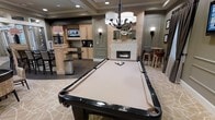 This single-floor entertainment lounge is thoughtfully designed to serve as a premier social and recreational space. At its center sits a full-size billiards table with dark wood rails and neutral felt, positioned beneath an elegant chandelier that anchors the room with warm, focused lighting. Wall sconces throughout the space complement the ambient light, contributing to a refined yet relaxed atmosphere.

A linear fireplace with a classic white mantel serves as a focal point along one wall, adorned with decorative accents that add a polished touch. Adjacent to the fireplace, a high-top bar seating area with stools provides an intimate spot for conversation.

On the opposite side, a well-appointed wet bar and kitchenette features light wood cabinetry, a raised countertop island, and a wall-mounted television — ideal for entertaining. Dark tile flooring in this zone transitions to a patterned carpet in the billiards area, subtly defining each functional space. The tray ceiling detail and carefully placed recessed lighting elevate the overall design, giving the lounge a finished, upscale character suited for community or private leisure use.