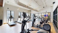 Located at 423 W Broad St in Columbus, Ohio, this well-appointed fitness center offers a spacious, single-floor layout designed to accommodate a full range of workout needs. The space features a thoughtfully designed interior that blends contemporary aesthetics with functional layout, making it suitable for both cardio and strength training.

The main cardio area is lined with large floor-to-ceiling windows that flood the space with natural light and provide views of the surrounding streetscape. Treadmills, ellipticals, and stationary bikes are arranged in open rows across light-toned hardwood flooring, creating an airy and energetic atmosphere. Striking starburst pendant light fixtures hang from the soaring ceilings, complementing the exposed ductwork for an industrial-modern design.

A dedicated free weights zone features tiered dumbbell racks, kettlebells, and open floor space for functional training. This area is distinguished by a warm wood slat ceiling, recessed lighting, and motivational text displayed on the wall, adding an inspiring ambiance. A power rack with a bench press station and adjustable benches support strength training, while floor-to-ceiling mirrors enhance the sense of space. Black-framed glass partitions divide sections of the gym while maintaining visual openness throughout.