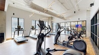 Located at 423 W Broad St in Columbus, Ohio, this well-appointed fitness center offers a spacious, single-floor layout designed to accommodate a full range of workout needs. The space features a thoughtfully designed interior that blends contemporary aesthetics with functional layout, making it suitable for both cardio and strength training.

The main cardio area is lined with large floor-to-ceiling windows that flood the space with natural light and provide views of the surrounding streetscape. Treadmills, ellipticals, and stationary bikes are arranged in open rows across light-toned hardwood flooring, creating an airy and energetic atmosphere. Striking starburst pendant light fixtures hang from the soaring ceilings, complementing the exposed ductwork for an industrial-modern design.

A dedicated free weights zone features tiered dumbbell racks, kettlebells, and open floor space for functional training. This area is distinguished by a warm wood slat ceiling, recessed lighting, and motivational text displayed on the wall, adding an inspiring ambiance. A power rack with a bench press station and adjustable benches support strength training, while floor-to-ceiling mirrors enhance the sense of space. Black-framed glass partitions divide sections of the gym while maintaining visual openness throughout.