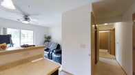 This single-floor residence offers a practical and comfortable layout designed for easy living. The living area is a welcoming space, featuring neutral white walls, carpeted flooring, and a ceiling fan that contributes to a relaxed atmosphere. Natural light filters in through a window, brightening the room and highlighting the cozy seating arrangement. A partial dividing wall separates the living area from an adjacent space, adding subtle definition to the open floor plan without sacrificing connectivity between rooms.

Warm wood-toned door frames and trim run throughout the interior, adding a touch of natural character to the home's clean, understated aesthetic. A hallway extends toward additional rooms, suggesting a well-organized layout that makes efficient use of the available space.

One of the standout features of this property is its serene outdoor setting. A front-facing deck or porch with a classic white railing overlooks a tranquil landscape of open green fields and mature deciduous trees, providing a peaceful and private outlook. The surrounding greenery creates a natural backdrop that enhances the overall sense of calm and openness. This home is well-suited for those seeking a quiet, low-maintenance living environment with a strong connection to the outdoors.