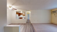 This single-floor residential unit offers a practical and well-organized layout designed for comfortable everyday living. The open living area serves as the heart of the space, featuring a combination of hardwood-style laminate flooring and plush wall-to-wall carpeting. The room is equipped with a wall-mounted air conditioning unit and a baseboard heating system, ensuring year-round climate control. Natural light enters through a window fitted with vertical blinds, brightening the neutral, cream-toned walls throughout.

The kitchen is positioned adjacent to the living area and features a pass-through opening that maintains an open, connected feel. Rich warm-toned wood cabinetry lines the walls, complemented by white appliances including a refrigerator, an electric range with overhead ventilation, and a dishwasher. A tile backsplash and laminate countertops complete the functional cooking space.

A hallway connects the living area to the bedroom, which is a generously sized, carpeted room with its own window dressed in vertical blinds and a baseboard heater beneath. The bedroom also includes a wall-mounted air conditioning unit, adding to the room's independent comfort. The overall ambiance of the unit is clean, neutral, and move-in ready.