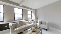 Situated in the heart of downtown Columbus, Ohio, this single-floor space at 50 W Broad St offers an elevated urban living experience with striking panoramic views of the city skyline. The interior is characterized by a refined, contemporary aesthetic, with a neutral color palette of crisp whites and soft grays that creates a clean, airy atmosphere throughout.

The living area is a standout feature, anchored by large black-framed windows that flood the space with natural light and frame impressive elevated views of the Columbus cityscape. The room is appointed with elegant tufted seating in ivory tones, accented by gold and glass furnishings that add a luxurious, sophisticated touch. Exposed structural beams along the ceiling contribute an architectural detail that complements the modern design sensibility.

The overall ambiance of the space is bright, open, and polished, blending classic elegance with contemporary urban style. Its prime downtown location provides easy access to Columbus's business district, dining, entertainment, and cultural destinations, making it an ideal setting for both professional and personal use.