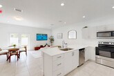 Located at 3031 NW Sixth St in Pompano Beach, FL, this two-story home offers a clean, modern aesthetic defined by crisp white walls, recessed lighting, and light-toned tile flooring throughout. The ground floor houses the main living spaces, including an open-plan living room, dining area, and kitchen. The kitchen is outfitted with white shaker cabinetry, light quartz countertops, a central island with sink, and a full suite of stainless steel appliances. The dining area adjoins the kitchen and benefits from natural light through a window overlooking the outdoor space. The living room is bright and airy, opening toward a balcony. A garage and entrance area complete the lower level. The upper floor contains two bedrooms, three bathrooms, a dedicated office, and generous closet space including walk-in configurations with wire shelving. The primary bedroom is well-proportioned and maintains the home's clean, minimalist palette. Bathrooms feature tile surrounds with tub-shower combinations. Two staircases with white railings and wood-toned treads connect the floors. Two balconies provide outdoor access, enhancing the overall sense of space and connection to the Florida surroundings.