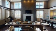 Located at 1252 Osgood St in North Andover, Massachusetts, this single-floor space serves as a sophisticated clubhouse or leasing center, thoughtfully designed to make a lasting impression. The centerpiece of the main lounge is a dramatic floor-to-ceiling dark stone fireplace surround, accented by a marble hearth and a mounted flat-screen television, flanked on either side by large multi-pane windows that flood the room with natural light. The soaring vaulted ceiling, finished with rich dark wood beams and coffered detailing, amplifies the grand sense of volume in the space. Wide-plank hardwood flooring flows throughout, grounding the open layout with warmth and continuity. The lounge area is furnished with plush sofas, accent chairs, and a rustic wood coffee table, creating an inviting social environment. A built-in nook features a tufted emerald velvet bench, patterned wallcovering, and curated artwork, offering an intimate seating alcove. Adjacent to the lounge, a kitchenette area is appointed with stone countertops, a geometric mosaic tile backsplash, dark cabinetry, a beverage refrigerator, and a water station. A workstation with a modern chair provides a functional area for leasing or administrative use. Together, these elements create a polished, welcoming atmosphere ideal for residents and visitors alike.