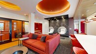 This dynamic single-floor clubroom is defined by its bold, energetic design aesthetic and diverse range of amenity spaces. The entrance is announced by a striking artistic mural wrapping the exterior entry doors, setting a creative tone throughout. Inside, the main lounge is anchored by a large orange sofa and complementary seating, beneath a dramatic tiered circular ceiling detail finished in warm copper tones. Suspended acrylic bubble chairs hang before a colorful letter-grid accent wall, adding a playful focal point. A dedicated "On Air" recording booth with glass walls provides a professional podcasting or broadcasting space. Built-in red upholstered nook seating offers quieter retreat areas alongside workstations framed by vivid starburst artwork. A separate media lounge features a wall-mounted television flanked by vertical wood slat paneling, generous grey sectional seating, and patterned stone-look tile flooring. The games area showcases a full-size billiards table beneath oversized metallic globe pendant lights, accompanied by a wet bar with built-in cabinetry and beverage refrigeration. A dedicated workspace near the exterior entrance provides a meeting table, red accent chairs, and access to printing equipment with views of the surrounding landscape outdoors.