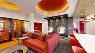 This dynamic single-floor clubroom is defined by its bold, energetic design aesthetic and diverse range of amenity spaces. The entrance is announced by a striking artistic mural wrapping the exterior entry doors, setting a creative tone throughout. Inside, the main lounge is anchored by a large orange sofa and complementary seating, beneath a dramatic tiered circular ceiling detail finished in warm copper tones. Suspended acrylic bubble chairs hang before a colorful letter-grid accent wall, adding a playful focal point. A dedicated "On Air" recording booth with glass walls provides a professional podcasting or broadcasting space. Built-in red upholstered nook seating offers quieter retreat areas alongside workstations framed by vivid starburst artwork. A separate media lounge features a wall-mounted television flanked by vertical wood slat paneling, generous grey sectional seating, and patterned stone-look tile flooring. The games area showcases a full-size billiards table beneath oversized metallic globe pendant lights, accompanied by a wet bar with built-in cabinetry and beverage refrigeration. A dedicated workspace near the exterior entrance provides a meeting table, red accent chairs, and access to printing equipment with views of the surrounding landscape outdoors.