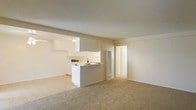 This single-story space features an open-concept design that seamlessly connects the living area and kitchen. The living area is finished with wall-to-wall neutral-toned carpet, creating a warm and understated foundation. A textured ceiling and soft, diffused lighting contribute to a calm, uniform ambiance throughout.

The kitchen is defined by a peninsula-style countertop that serves as a natural divider between the two spaces while maintaining visual openness. White cabinetry lines the kitchen walls, offering ample storage, while the countertop provides functional workspace. A sink is positioned along the peninsula, and the clean white finishes give the kitchen a bright, tidy appearance.

A hallway visible from the main living area leads to additional rooms, suggesting a practical and straightforward floor plan. The overall color palette is consistently neutral — whites, beiges, and light grays — lending the space a clean, move-in-ready aesthetic. The open layout and connected spaces make the interior feel cohesive and easy to navigate, suitable for a variety of furnishing arrangements.