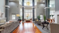 This elegantly designed single-floor community space makes an immediate impression with its dramatic double-height ceilings and stately dark columns that frame a sweeping curved bay window, flooding the main lounge with abundant natural light and views of the surrounding grounds. Rich hardwood flooring flows seamlessly throughout, grounding the space in warmth and sophistication.

The main lounge area is thoughtfully appointed with multiple seating arrangements, including plush sofas, houndstooth-patterned accent chairs, and classic area rugs, all centered around a beautifully crafted white fireplace mantel with an elegant surround. A large statement pendant light anchors the soaring ceiling above, enhancing the grand scale of the room.

Adjacent to the lounge, a dedicated billiards room offers a full-size pool table set beneath another soaring ceiling, accompanied by its own fireplace — making it an ideal space for recreation and social gathering. Tall interior windows with white millwork connect this room to the wider space while maintaining a sense of defined purpose.

The kitchenette features rich cherry wood cabinetry, light stone countertops, a breakfast bar with seating, patterned wallcovering, and a full-size black refrigerator — well-suited for hosting events. Throughout the space, lush greenery, curated artwork, and warm accent lighting contribute to a refined yet welcoming ambiance.