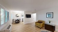 Located at 45 Tamal Vista Blvd in Corte Madera, CA, this single-floor, two-bedroom, one-bathroom residence offers a clean, modern aesthetic throughout. Light wood flooring flows seamlessly across every room, lending a bright and cohesive feel to the entire space. The open-concept living area is generously proportioned, featuring a wall-mounted flat-screen TV, ample room for seating, and a smooth transition into the dining area. The kitchen is efficiently designed with white cabinetry, quartz countertops, a subway tile backsplash, and industrial-style open shelving, creating a functional yet stylish workspace. The spacious primary bedroom benefits from natural light through plantation-shuttered windows, a mirrored sliding closet, and wall-mounted bedside lighting. The second bedroom is equally well-lit, featuring additional plantation shutters and its own closet. A hallway connects the bedrooms to three closets for ample storage. The full bathroom is finished with classic white subway tile surrounding the shower/tub combo, complemented by a granite-topped vanity and chrome fixtures. Crisp white walls and understated décor throughout contribute to the home's calm and welcoming ambiance.