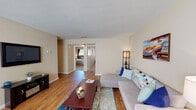 This single-floor, two-bedroom, one-bathroom residence offers a comfortable and functional layout designed to maximize everyday living. The open living area serves as the heart of the home, featuring warm hardwood-style flooring, neutral beige walls, and a ceiling fan that contributes to a light and airy atmosphere. A wall-mounted flat-screen TV and ample space for seating make it an inviting area for relaxation. The living space flows naturally toward a dining nook positioned near a large window with blinds, allowing abundant natural light to fill the area. The kitchen is equipped with warm oak cabinetry, granite-style countertops, a double stainless steel sink, and a dishwasher, providing a practical and attractive workspace. A full-size refrigerator completes the kitchen's functionality. The bedrooms feature plush carpeting and windows that allow natural light in, with the primary bedroom offering room for a full suite of furniture. The shared full bathroom includes a clean, white tub-shower combination. Throughout the residence, the consistent use of hardwood-style flooring in common areas and soft carpet in the bedrooms creates a cohesive, comfortable feel well-suited for everyday living.