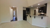 This two-floor home is designed with an open and functional layout that maximizes both space and comfort. The main living area immediately impresses with its soaring vaulted ceilings, which lend a grand sense of airiness to the space. Soft, neutral-toned carpet covers the living area floor, creating a warm and inviting atmosphere, while dark hardwood flooring runs through the adjoining hallway and entryway, providing a rich visual contrast.

The kitchen is a standout feature, showcasing bold dark cabinetry that creates a striking visual contrast against the light walls. A stainless steel refrigerator anchors the culinary space, which also includes a peninsula-style breakfast bar with a granite-look countertop and bar seating — ideal for casual dining or entertaining. Recessed lighting throughout ensures the space is well-lit and functional.

A wide hallway connects the living area to the front entryway, where a paneled front door with sidelights allows natural light to filter in. The layout flows naturally between spaces, with multiple doorways offering clear sightlines and easy accessibility throughout. The overall design combines practical functionality with clean, modern finishes, making this a well-appointed and comfortable multi-level home.