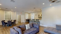 This well-appointed clubroom in Fairfax, Virginia, offers a thoughtfully designed single-floor layout with distinct functional zones, all unified by warm hardwood flooring, neutral tones, and abundant natural light. The living area provides a comfortable lounge setting with plush seating, a wall-mounted flat-screen TV, recessed lighting, and an elegant chandelier, creating an inviting space for relaxation or gatherings. French doors connect the living area to adjacent spaces, enhancing the open flow throughout. The main dining area features soaring vaulted ceilings, large windows with lush outdoor views, and bistro-style high-top tables, while a secondary dining nook offers tufted seating around a round table alongside a dark granite countertop bar. The kitchen is a standout, showcasing a curved granite island, dark cabinetry with glass-front uppers, a striking mosaic tile backsplash in earth tones, stainless steel appliances, and travertine tile flooring. A dedicated workspace is lined with a long built-in desk beneath floor-to-ceiling windows that flood the room with natural light and scenic greenery views. Throughout, the space balances practicality with refined design, making it a versatile and welcoming community facility.