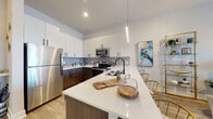 This thoughtfully designed single-floor residence blends contemporary style with functional living across several well-appointed spaces. The open-concept kitchen is a standout feature, showcasing a two-tone cabinet design with crisp white upper cabinets and rich dark wood lower cabinets, complemented by white quartz countertops and a full suite of stainless steel appliances. A generous island with pendant lighting serves as both a prep space and casual dining area, with seating for multiple guests. Light wood-style flooring flows throughout the main living areas, enhancing the bright and airy atmosphere. The living room offers a comfortable, welcoming ambiance, bathed in natural light from large windows fitted with sheer curtains that provide a soft, diffused glow. The bedroom continues the theme of natural light, featuring expansive windows with an outdoor view and a bold accent wall that adds a distinctive pop of color. The bathroom is clean and modern, finished with wood-look tile flooring, a sleek vanity, and a tub-shower combination with matte black fixtures. A dedicated home office or den rounds out the space, distinguished by a striking geometric accent wall and built-in shelving, offering a creative and productive retreat within the residence.