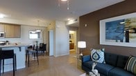 This single-floor residence offers a well-designed open-concept layout that seamlessly connects the living, dining, and kitchen areas. The living space is anchored by a bold accent wall in a rich espresso brown tone, creating a striking focal point that complements the neutral warm beige palette found throughout the home. Large-format tile flooring runs consistently across the main living areas, lending a clean and cohesive look underfoot. The living area is bright and comfortable, featuring a contemporary sofa accented with decorative pillows and a glass-topped coffee table that adds an elegant, airy touch. A modern floor lamp provides warm ambient lighting. Adjacent to the living space, the dining area accommodates a dark-framed table and chairs beneath a pendant light fixture, enhancing the intimate dining experience. Natural light filters in through a window nearby, brightening the space. Down the hallway, views into what appears to be a bathroom and additional rooms suggest a practical and efficiently organized floor plan. The kitchen cabinetry is visible in the background, offering functional storage and workspace. Overall, the space delivers a modern residential aesthetic with a warm, welcoming ambiance suited for comfortable everyday living.