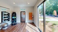 This single-floor yoga and fitness studio is thoughtfully designed to foster a calm, focused environment for movement and mindfulness. The space features warm hardwood flooring throughout, providing both an aesthetic appeal and a practical surface for a variety of fitness activities. Large sliding glass doors and expansive windows line one wall, flooding the studio with natural light and offering views of the surrounding green landscape, creating a connection to the outdoors that enhances the sense of openness and tranquility. A full-length wall mirror reflects the room, visually expanding the space and allowing practitioners to monitor their form during sessions. Vibrant orange yoga mats are neatly stored on a wall-mounted wooden rack, accompanied by a conveniently placed hand sanitizer station. Exercise balls and a selection of weighted equipment are organized on nearby storage racks, keeping the studio functional and orderly. Bold, orange circular wall graphics display motivational phrases such as "I bend so I don't break" and "The quieter you become, the more you are able to hear," reinforcing the studio's wellness-focused philosophy. A teal "Namaste" script above the entrance door adds a welcoming, mindful touch. Recessed ceiling lighting ensures the space remains bright and inviting at all hours.