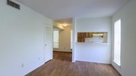 This single-floor residence offers a clean, functional layout designed for comfortable everyday living. The interior is unified by warm hardwood-style flooring that flows throughout the main living areas, lending a cohesive and inviting feel to the space. Crisp white walls provide a neutral backdrop that enhances the sense of openness and light. A pass-through kitchen counter connects the kitchen to the adjacent living area, promoting an open, connected flow between spaces. The kitchen is fitted with warm-toned wood cabinetry and includes a range hood, offering practical storage and functionality. A louvered closet door adds a classic architectural touch near the entryway, providing convenient storage access. Natural light filters in through windows dressed with white blinds, brightening the interior throughout the day. The layout is straightforward and efficient, with clear sightlines from the main living area into the adjoining rooms. Ceiling-mounted lighting provides additional illumination in the transitional spaces. Overall, the space presents a tidy, well-maintained environment with a neutral palette and practical design elements suited for a variety of living arrangements.