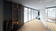 This single-floor yoga and fitness studio offers a thoughtfully designed space for both mindful movement and structured exercise. The main yoga area is characterized by a distinctive striped wood-effect floor that runs the length of the room, creating a visually dynamic foundation. Floor-to-ceiling glass windows along one wall flood the space with natural light while providing views of surrounding greenery, contributing to a calm and open atmosphere. Full-length mirrored walls expand the sense of space and allow practitioners to monitor their form, while a sleek wall-mounted ballet barre adds a dedicated element for barre and stretching exercises. A wooden wall-mounted ladder bar with TRX suspension straps provides additional functional training options. Stability balls are neatly stored along the windowed corridor, keeping the main floor clear and uncluttered. Adjacent to the yoga studio, a well-equipped fitness area houses a variety of cardio machines, cable machines, and strength equipment, along with a rack of kettlebells and dumbbells. Recessed ceiling lighting throughout maintains a clean, modern aesthetic, while blue-grey accent walls add depth and a focused, energizing ambiance to the overall space.