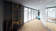 This single-floor yoga and fitness studio offers a thoughtfully designed space for both mindful movement and structured exercise. The main yoga area is characterized by a distinctive striped wood-effect floor that runs the length of the room, creating a visually dynamic foundation. Floor-to-ceiling glass windows along one wall flood the space with natural light while providing views of surrounding greenery, contributing to a calm and open atmosphere. Full-length mirrored walls expand the sense of space and allow practitioners to monitor their form, while a sleek wall-mounted ballet barre adds a dedicated element for barre and stretching exercises. A wooden wall-mounted ladder bar with TRX suspension straps provides additional functional training options. Stability balls are neatly stored along the windowed corridor, keeping the main floor clear and uncluttered. Adjacent to the yoga studio, a well-equipped fitness area houses a variety of cardio machines, cable machines, and strength equipment, along with a rack of kettlebells and dumbbells. Recessed ceiling lighting throughout maintains a clean, modern aesthetic, while blue-grey accent walls add depth and a focused, energizing ambiance to the overall space.