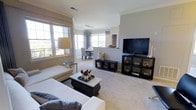 This single-floor, two-bedroom, two-bathroom residence offers an open and well-connected floor plan designed for comfortable everyday living. The living area is carpeted and generously proportioned, accommodating a large sectional sofa, a stylish coffee table, and an entertainment center, creating a cohesive and functional gathering space. Large windows along multiple walls flood the interior with abundant natural light, enhancing the bright, airy atmosphere throughout.

Crown molding adds a refined architectural detail to the living space, while track lighting on the ceiling provides flexible illumination options. The living area transitions smoothly into the dining space, which is positioned near a corner of windows, offering pleasant outdoor views. A half-wall with a pass-through opening connects the dining area to the kitchen, maintaining an open feel while providing subtle spatial definition between the two areas.

The neutral color palette—featuring soft gray walls and light-toned carpet—creates a clean, versatile backdrop throughout the home. The overall ambiance is warm and inviting, with natural light and thoughtful layout details making the space feel comfortable and move-in ready.