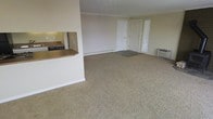 This single-floor residential unit offers a straightforward and functional layout designed for comfortable everyday living. The main living area is generously sized, finished with wall-to-wall carpeting in a neutral beige tone that extends throughout the space and contributes to a warm, understated atmosphere. The walls are painted in a light cream color, keeping the interior feeling bright and open.

A notable architectural feature is the pass-through kitchen window, complete with a wooden countertop overhang that connects the kitchen to the living area. This design element adds convenience and openness, allowing interaction between the two spaces. A glimpse into the kitchen reveals dark appliances and additional counter space.

Natural light enters the living area through a slatted window blind, adding a modest level of brightness to the room. A wall-mounted HVAC unit is positioned along one wall, providing climate control. Baseboard heating is also visible, offering supplemental warmth. Two interior doors lead to additional areas of the unit, suggesting the presence of a bedroom and a secondary room or closet space.

Overall, the unit presents a clean, neutral palette with practical features throughout, making it a functional and move-in ready residential space.